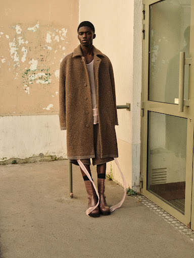 Fashion shot of a Black man in a tan coat and boots, standing in front of a weathered wall and doorway with ribbons attached to his legs.