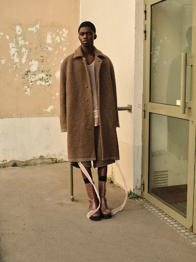 Fashion shot of a Black man in a tan coat and boots, standing in front of a weathered wall and doorway with ribbons attached to his legs.