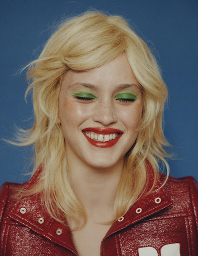 Smiling woman with blonde hair, green eyeliner and red lipstick posing in front of a blue background, wearing a red jacket.