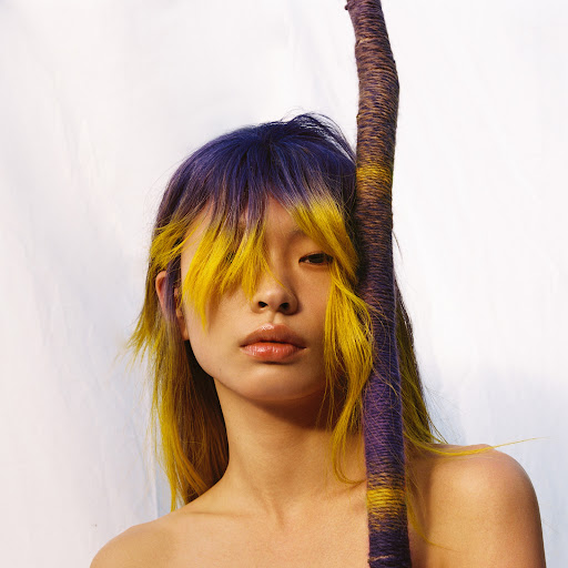 Portrait of an Asian woman with purple and yellow hair posing with a branch wrapped in purple and yellow yarn against a white backdrop.