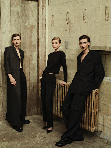 Three fashion models strike poses in a studio against a concrete and wood wall. Two male models and one female model wear dark clothing.