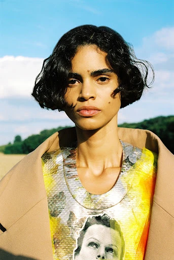 Woman in a printed dress and coat stands outdoors against a blue sky. Fashion shot in a natural setting with soft light.