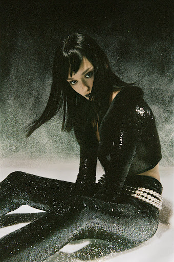 Adult female with dark hair and dark lipstick poses in a shiny black ensemble. She sits in a studio against a grey background.