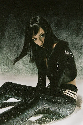 Adult female with dark hair and dark lipstick poses in a shiny black ensemble. She sits in a studio against a grey background.