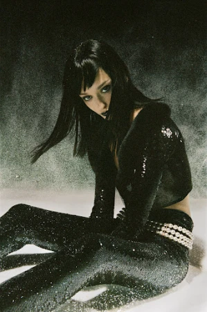 Adult female with dark hair and dark lipstick poses in a shiny black ensemble. She sits in a studio against a grey background.