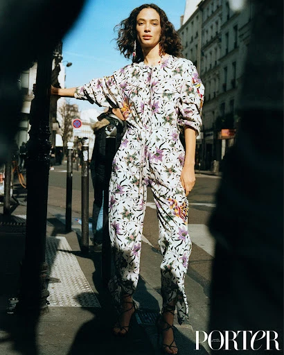 Fashion editorial shot of a model in a floral jumpsuit on a city street, posing near a pole with buildings in the background.