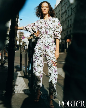 Fashion editorial shot of a model in a floral jumpsuit on a city street, posing near a pole with buildings in the background.