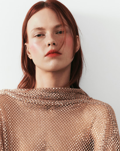 Close-up studio portrait of a woman with auburn hair wearing a sparkling bronze net top against a white background.