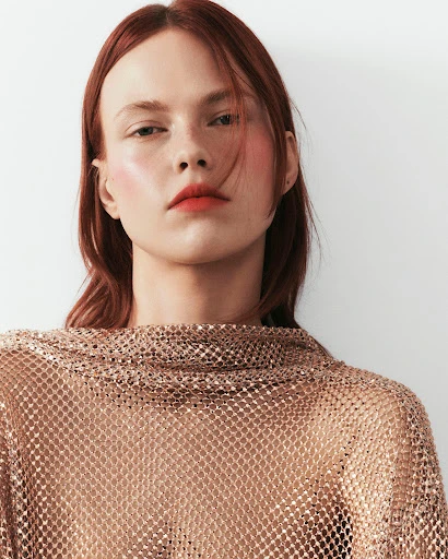 Close-up studio portrait of a woman with auburn hair wearing a sparkling bronze net top against a white background.
