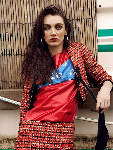 A woman poses in a patterned jacket and skirt against a textured backdrop, featuring bold red and blue accents with soft, diffused lighting.