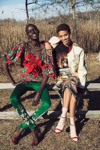 Two smiling women pose outdoors, one in a floral blouse and green pants, the other in a graphic tee and printed skirt, sunny and bright.