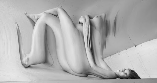 Black and white abstract studio portrait of a nude woman with stretched features, lying on her side with a concrete floor.