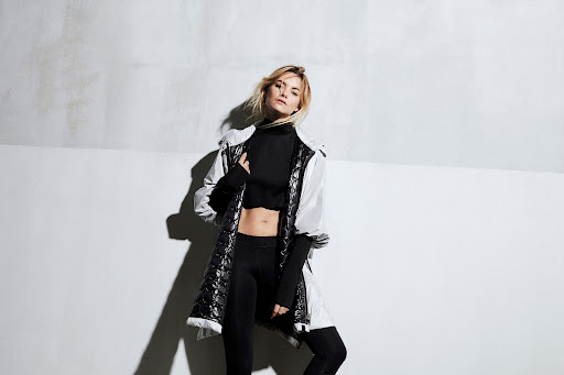 Stylish blonde woman in black top and leggings, wearing a white jacket, poses in a modern indoor studio with a textured white wall.