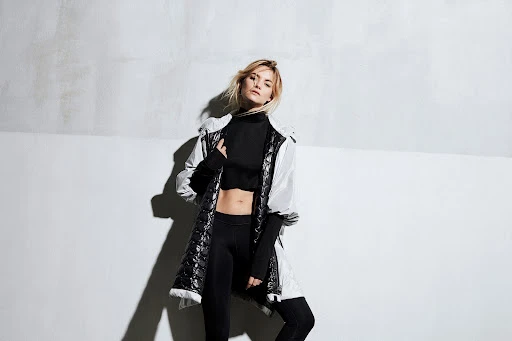 Stylish blonde woman in black top and leggings, wearing a white jacket, poses in a modern indoor studio with a textured white wall.