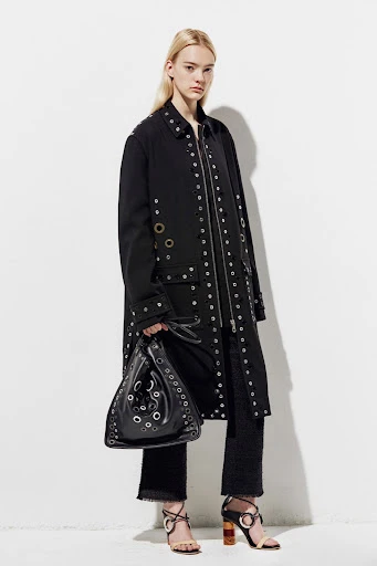 Fashion shot of a fair-skinned woman in a black jacket with silver eyelets, holding a matching black eyeletted purse against a white wall.