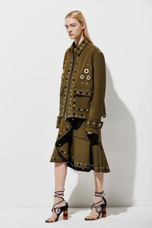 Model wearing an olive green grommet-embellished jacket and skirt with lace-up sandals against a plain white background