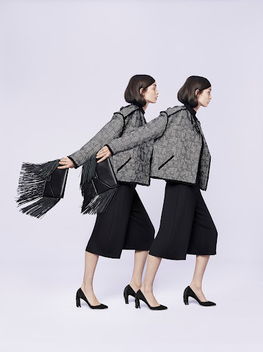 Two fashion models in tweed jackets and black culottes walk in sync against a neutral background, one carrying a fringe bag.