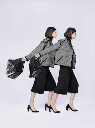 Two fashion models in tweed jackets and black culottes walk in sync against a neutral background, one carrying a fringe bag.
