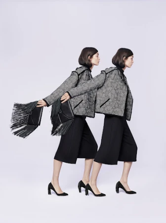 Two fashion models in tweed jackets and black culottes walk in sync against a neutral background, one carrying a fringe bag.
