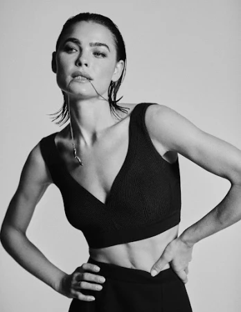 Black and white portrait of a woman with slicked-back wet hair holding a thin necklace chain in her mouth against a plain background.
