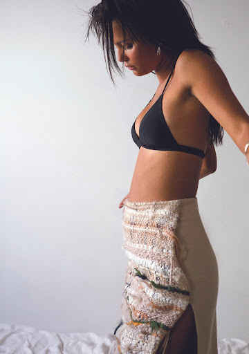 Woman in a black bikini top and beige skirt with lace detail poses indoors against a white background, looking down, in soft lighting.