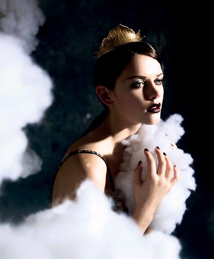 A woman wearing a gold crown and dark lipstick posing among white fluffy cloud props in a dark studio setting.