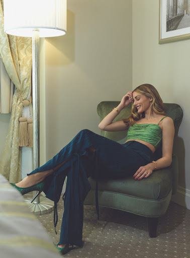 Blonde woman poses in green top and blue pants, sitting in a chair near a lamp in an indoor setting, fashion editorial shot.