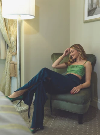 Blonde woman poses in green top and blue pants, sitting in a chair near a lamp in an indoor setting, fashion editorial shot.