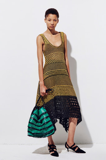 A single young adult female fashion model in a yellow and black knit dress poses in front of a white wall, holding a striped bag.