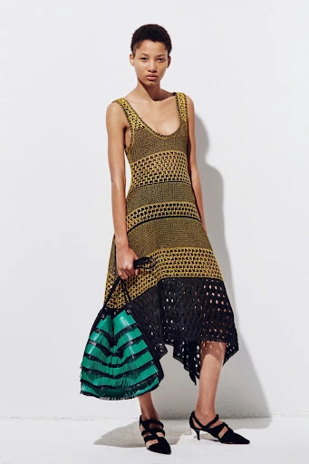 A single young adult female fashion model in a yellow and black knit dress poses in front of a white wall, holding a striped bag.
