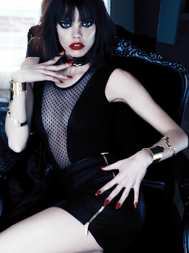 Dark-haired woman with red lipstick sits in a black chair wearing a black outfit and gold jewelry, hands near her face.