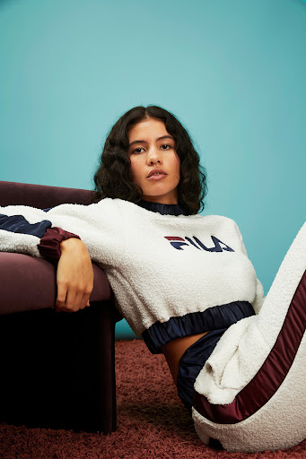 A woman with wavy dark hair sits on a plush pink carpet, wearing a white and navy blue FILA fleece tracksuit, against a blue backdrop.