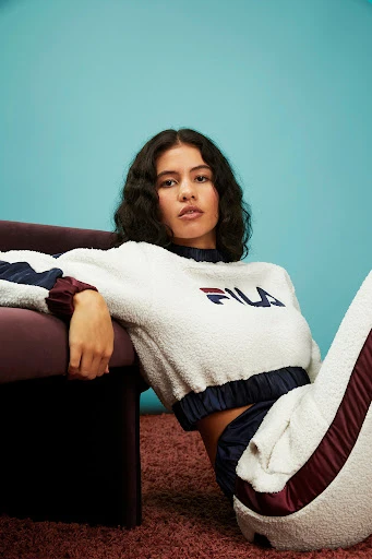 A woman with wavy dark hair sits on a plush pink carpet, wearing a white and navy blue FILA fleece tracksuit, against a blue backdrop.