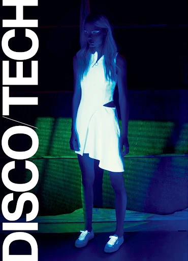 A woman with blonde hair wearing a white dress and white sneakers stands indoors in front of a green and black background.