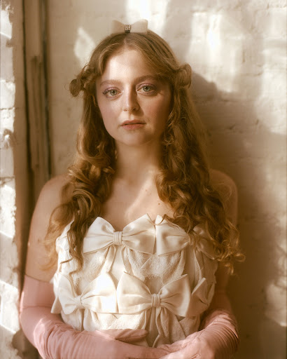 Portrait of a curly-haired young woman with a neutral expression wearing a cream-colored dress with bows in an indoor setting with window lighting.