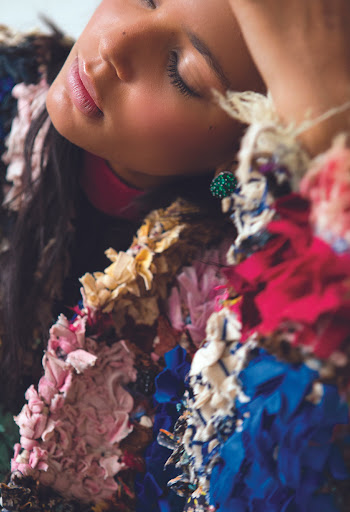 Close-up of a woman with fair skin and dark hair, eyes closed, wearing a colorful ruffled garment, portrait orientation.