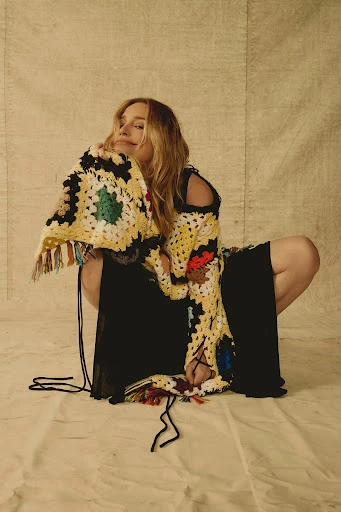 Woman in patterned sweater and black dress poses against neutral backdrop in portrait shot, fashion editorial.