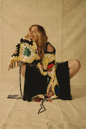 Woman in patterned sweater and black dress poses against neutral backdrop in portrait shot, fashion editorial.