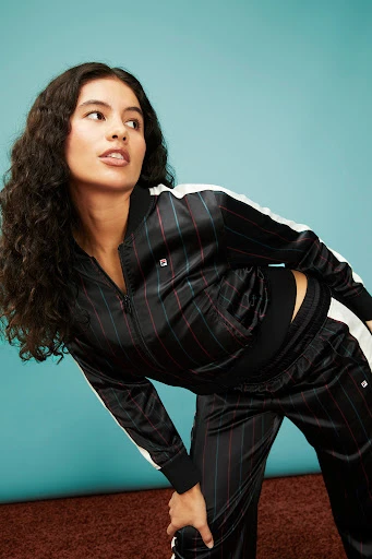 Portrait of a woman in a striped black tracksuit with white stripes on a brown rug in front of a blue wall, looking off-camera.