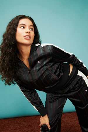 Portrait of a woman in a striped black tracksuit with white stripes on a brown rug in front of a blue wall, looking off-camera.