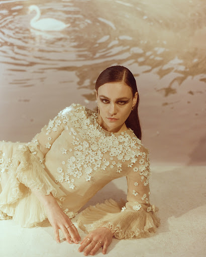 Brunette woman in a floral dress lies in front of a faded backdrop with a swan image, creating an ethereal and artistic fashion portrait.
