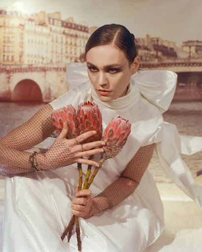 Elegant woman in white dress poses holding three protea flowers against a Parisian backdrop, displaying high fashion and editorial style.