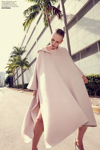 Fashion shot of an adult female model posing in a beige poncho-style coat with palm trees and an urban building in the background.