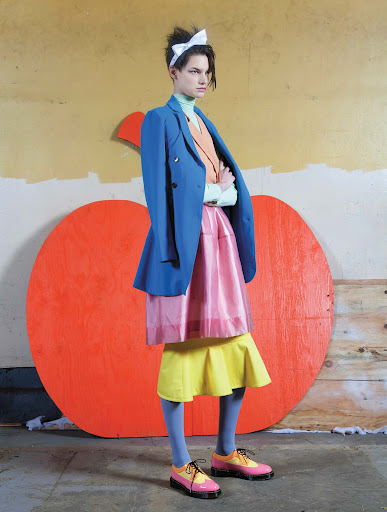 A fashion photo of a person with arms crossed, wearing a blue blazer and layered skirts, posing in front of a painted apple.