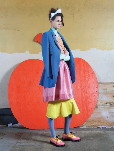 A fashion photo of a person with arms crossed, wearing a blue blazer and layered skirts, posing in front of a painted apple.
