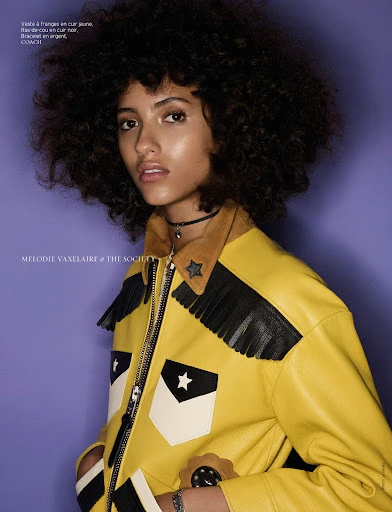 Close-up portrait of a woman with curly dark hair, wearing a yellow leather jacket with black fringe, posing against a solid purple backdrop.