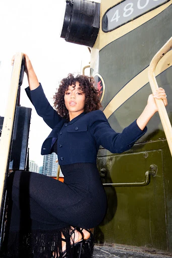 Woman posing on a train wearing a dark blue cropped jacket and long black skirt with a city skyline in the background.