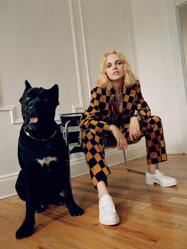 Blonde woman in checkered suit sits next to a black dog indoors on a hardwood floor in front of white decorative wall panels.