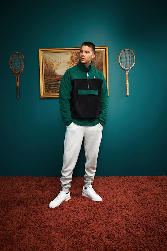 Man posing indoors wearing a black and green fleece jacket, sweatpants, and sneakers. Behind him hang tennis rackets and a painting.