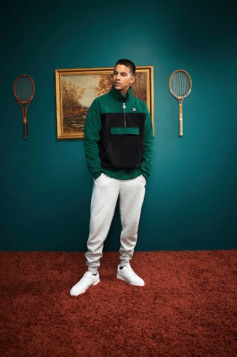 Man posing indoors wearing a black and green fleece jacket, sweatpants, and sneakers. Behind him hang tennis rackets and a painting.